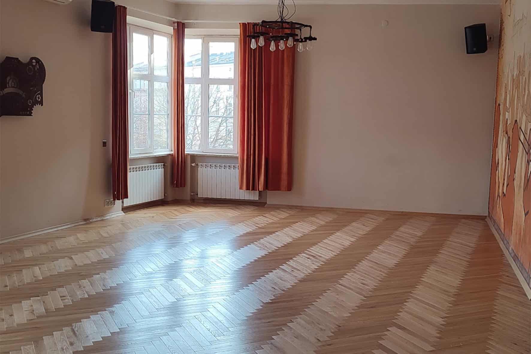 Duży pokój z drewnianym parkietem i dużymi oknami z zasłonami.